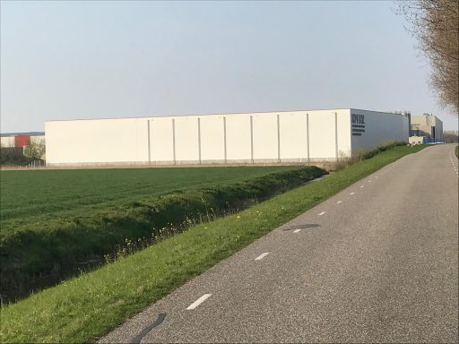 Een lang, wit gebouw naast een groene akker langs een asfaltweg.